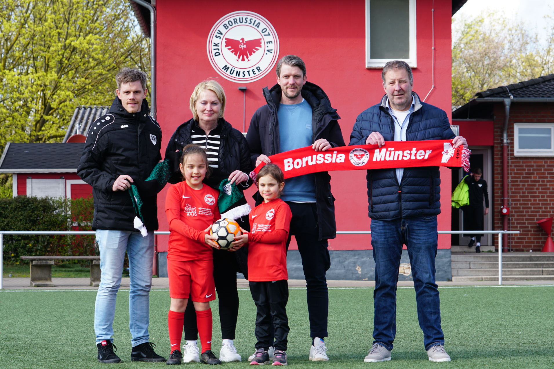 F&uuml;nf Personen stehen auf einem Fu&szlig;ballplatz, halten einen roten Schal mit &bdquo;Borussia M&uuml;nster&ldquo; und einen Fu&szlig;ball.
