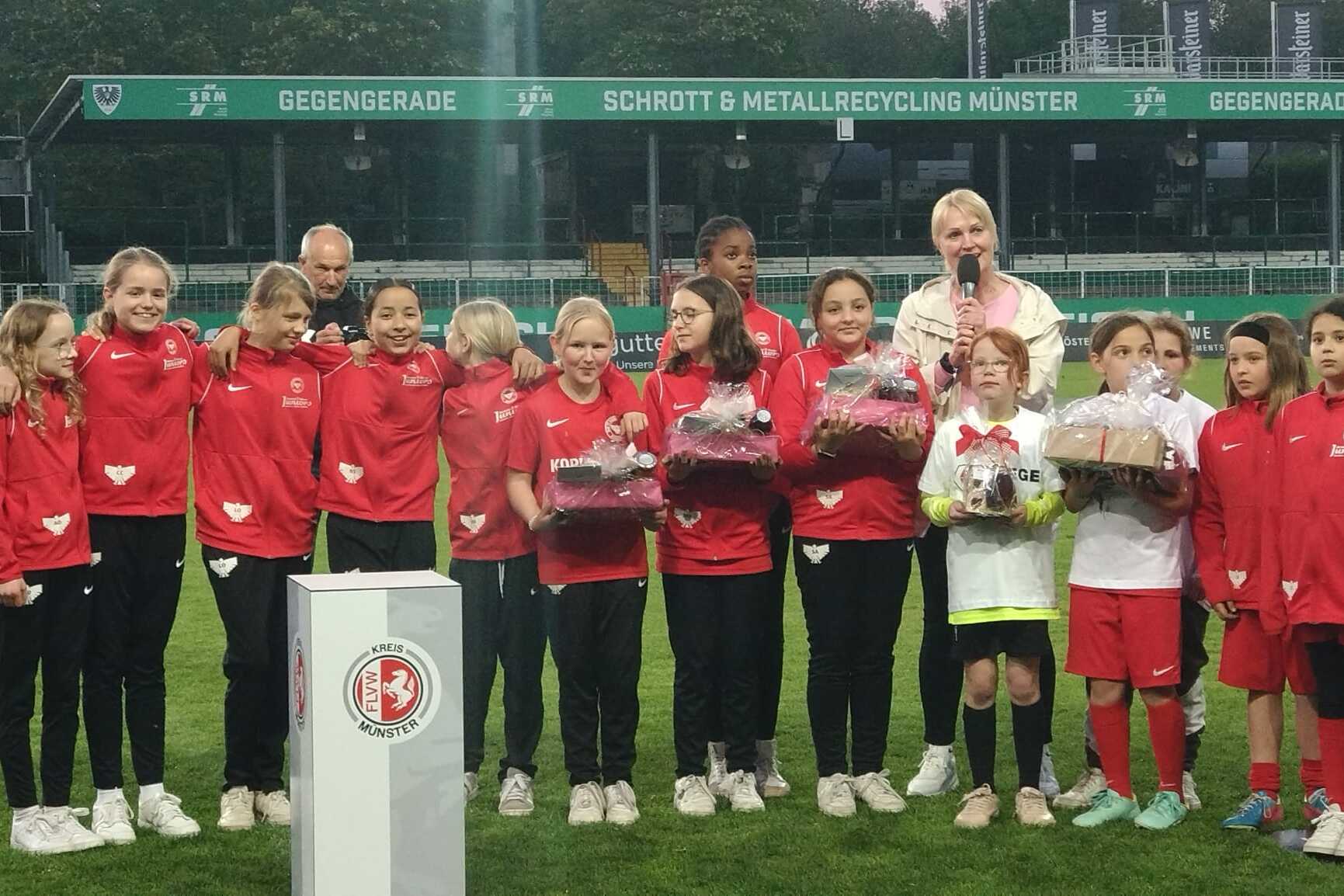 M&auml;dchenmannschaft in roten Trainingsanz&uuml;gen sammelt sich auf dem Spielfeld f&uuml;r eine Ehrung mit Pr&auml;sentk&ouml;rben.