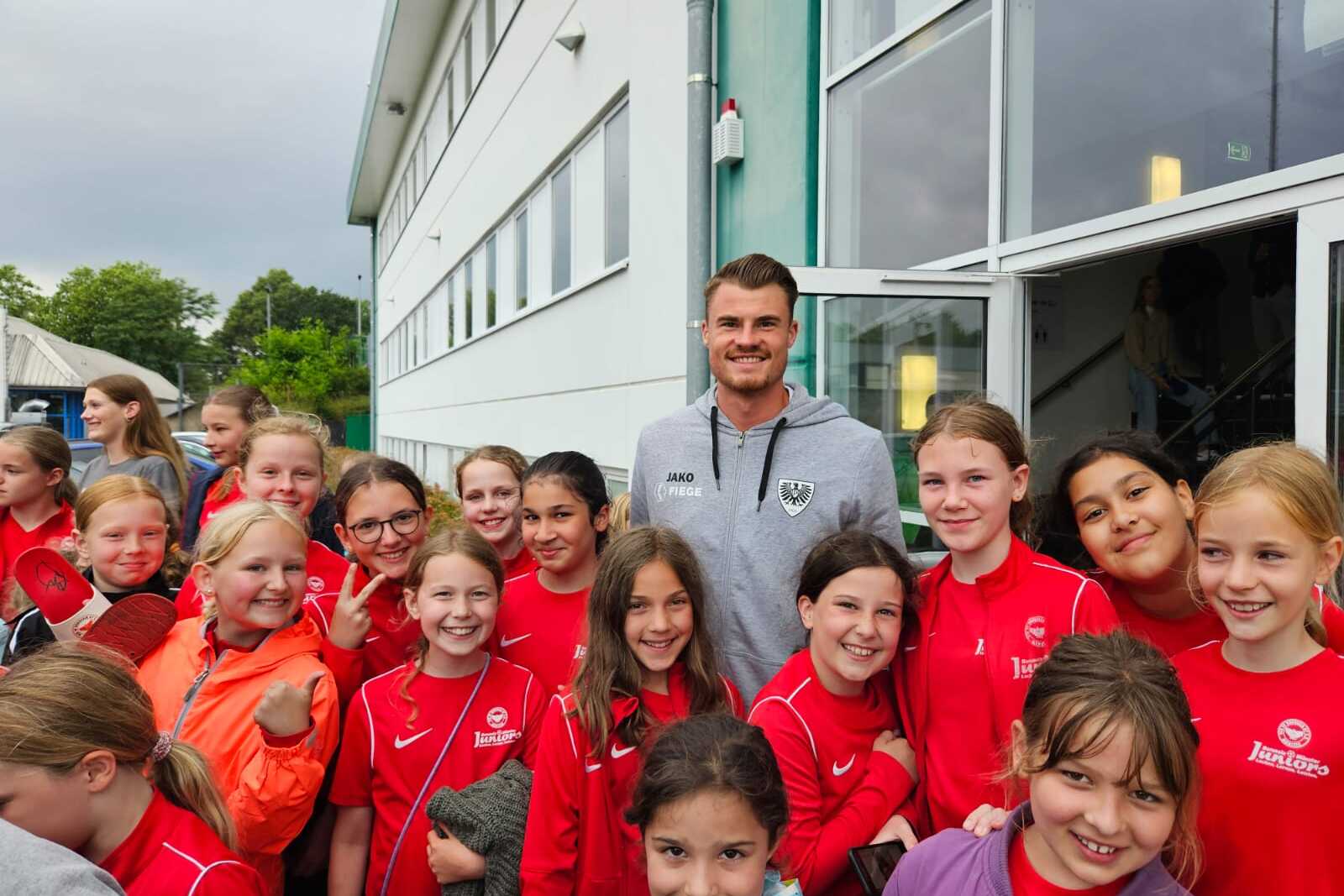 Gruppe von M&auml;dchen in roten Sportjacken posiert mit einem l&auml;chelnden Trainer vor einem Veranstaltungsgeb&auml;ude.