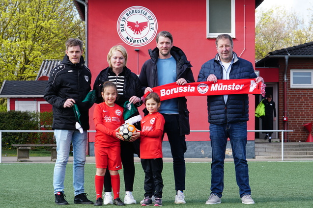 F&uuml;nf Personen stehen auf einem Fu&szlig;ballplatz, halten einen roten Schal mit &bdquo;Borussia M&uuml;nster&ldquo; und einen Fu&szlig;ball.