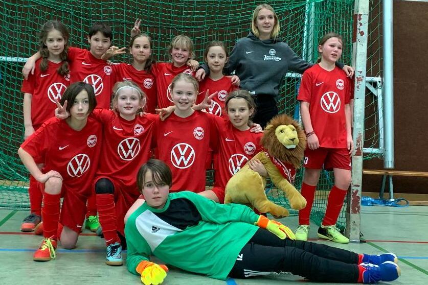 M&auml;dchenfu&szlig;ballmannschaft in roten Trikots posiert vor einem Tor, eine Spielerin in Torwartuniform liegt vorn mit einem Pl&uuml;schl&ouml;wen.