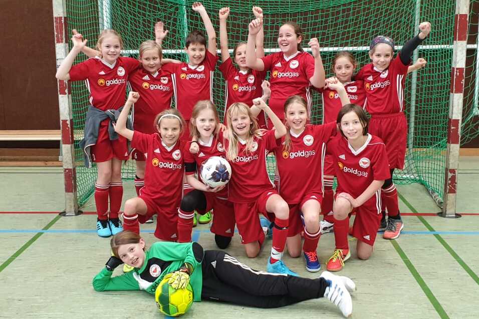 Gruppentraining einer M&auml;dchenequipe in roten Fu&szlig;balltrikots, posierend vor einem Tor mit B&auml;llen und Jubelgesten.