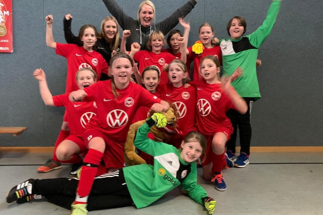 M&auml;dchenfu&szlig;ballmannschaft in roten Trikots posiert fr&ouml;hlich nach dem Spiel, einige halten Torwarthandschuhe und feiern.