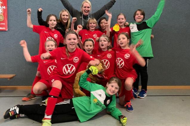 M&auml;dchenfu&szlig;ballmannschaft in roten Trikots posiert fr&ouml;hlich nach dem Spiel, einige halten Torwarthandschuhe und feiern.