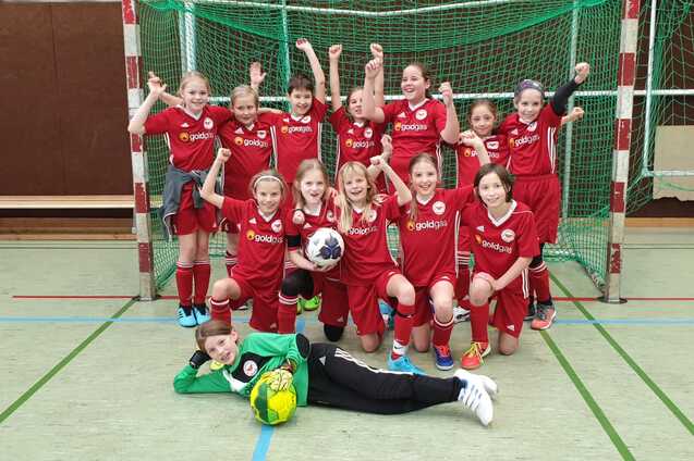 Gruppentraining einer M&auml;dchenequipe in roten Fu&szlig;balltrikots, posierend vor einem Tor mit B&auml;llen und Jubelgesten.