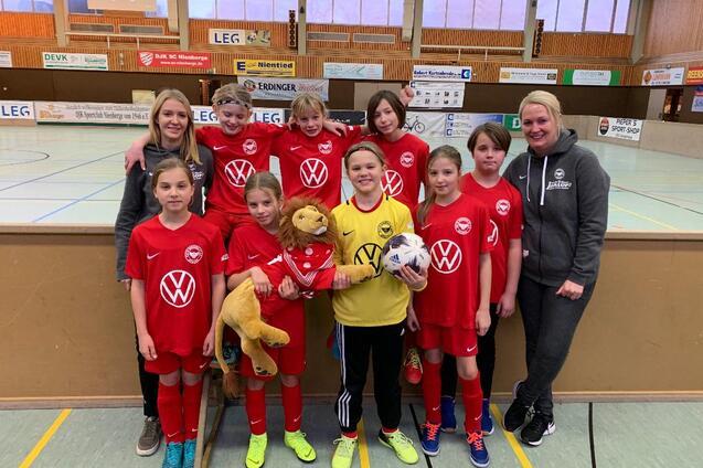 M&auml;dchenfu&szlig;ballmannschaft in roten Trikots mit einer Puppe und einem Ball, umrahmt von Trainerinnen in einer Sporthalle.