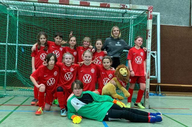 M&auml;dchenfu&szlig;ballmannschaft in roten Trikots posiert vor einem Tor, eine Spielerin in Torwartuniform liegt vorn mit einem Pl&uuml;schl&ouml;wen.