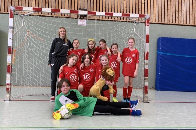 Gruppe von M&auml;dchen in roten Fu&szlig;balltrikots steht vor einem Tor, eine M&auml;dchen tr&auml;gt ein L&ouml;wenkost&uuml;m, torwart liegt im Vordergrund.