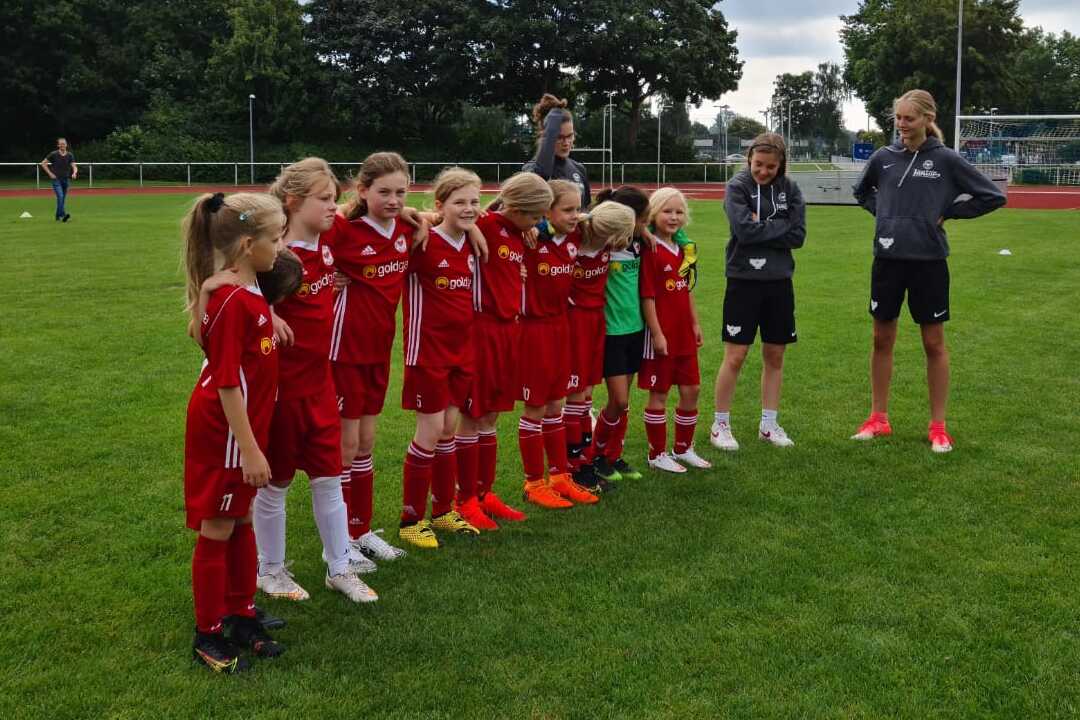 Gruppe junger Fu&szlig;ballspielerinnen in roten Trikots steht auf einem Rasenplatz, beobachtet von Trainern.
