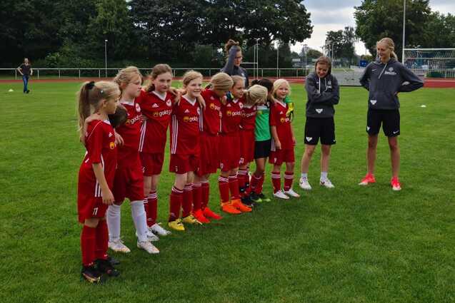 Gruppe junger Fu&szlig;ballspielerinnen in roten Trikots steht auf einem Rasenplatz, beobachtet von Trainern.