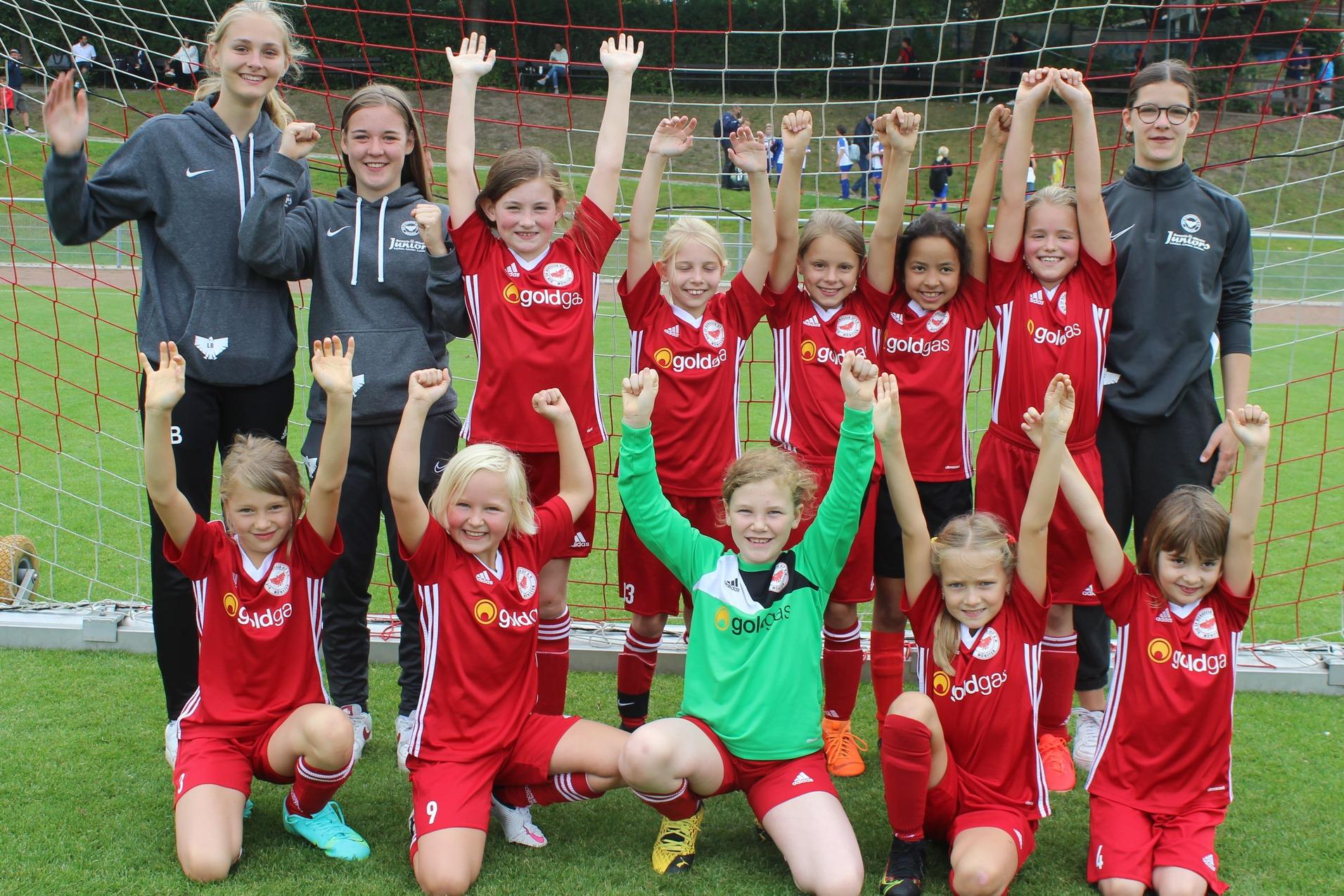 M&auml;dchenfu&szlig;ballteam in roten Trikots posiert fr&ouml;hlich vor einem Fu&szlig;balltor, einige Spielerinnen heben die Arme.