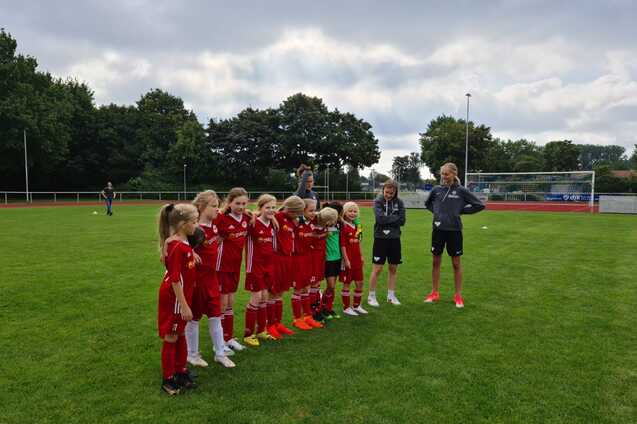 Gruppe junger Fu&szlig;ballspielerinnen in roten Trikots steht auf einem Rasenplatz, beobachtet von Trainern.