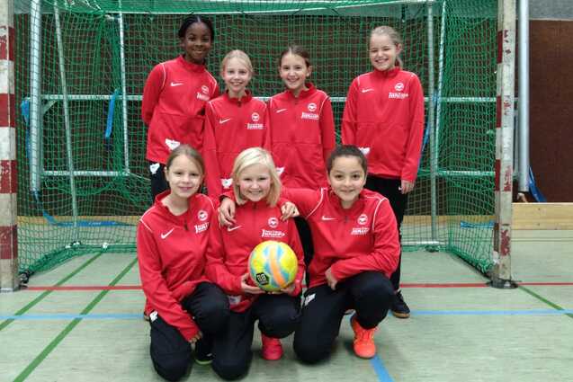 Gruppe von sieben M&auml;dchen in roten Trainingsanz&uuml;gen, posierend vor einem Handballtor mit einem Ball in der Mitte.