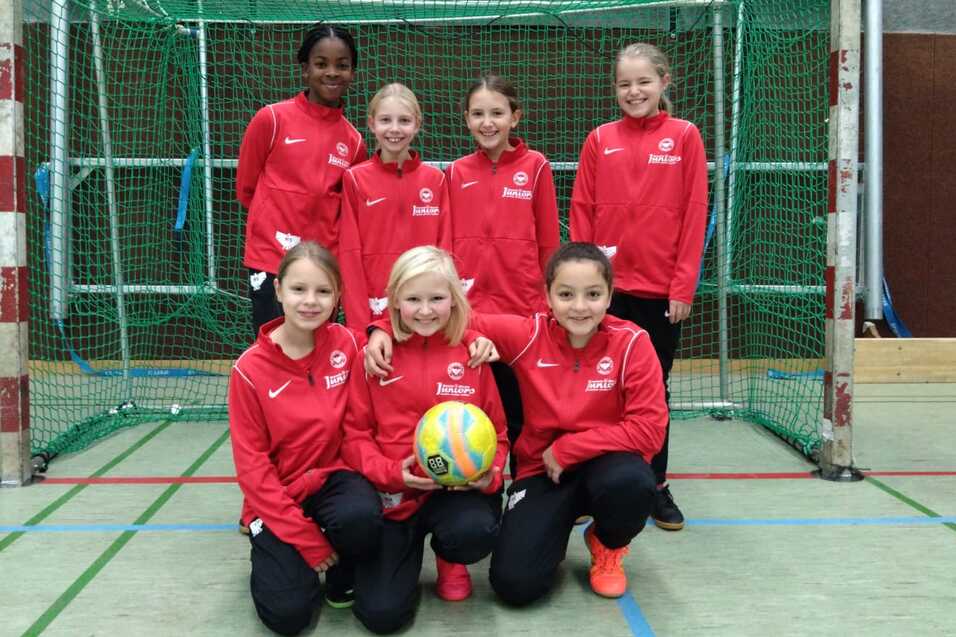 Gruppe von sieben M&auml;dchen in roten Trainingsanz&uuml;gen, posierend vor einem Handballtor mit einem Ball in der Mitte.