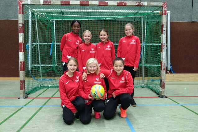 Gruppe von sieben M&auml;dchen in roten Trainingsanz&uuml;gen, posierend vor einem Handballtor mit einem Ball in der Mitte.