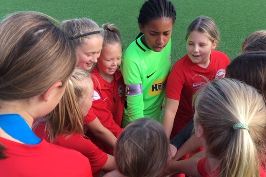 M&auml;dchenfu&szlig;ballmannschaft in roten Trikots versammelt sich um die Torh&uuml;terin in gr&uuml;nem Trikot f&uuml;r einen Teamtreff.