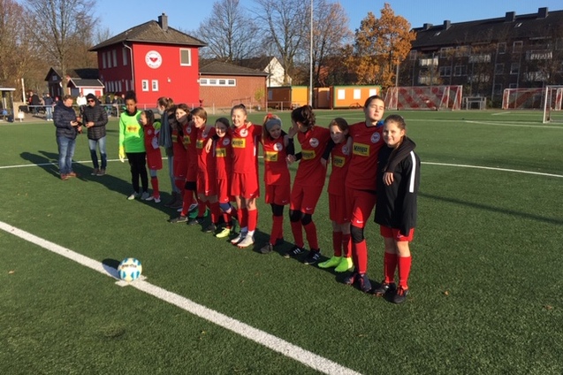 M&auml;dchen-Fu&szlig;ballmannschaft in roten Trikots steht auf einem Kunstrasenplatz, bereit f&uuml;r das Spiel. Ein Ball liegt vor ihnen.