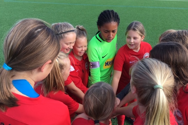 M&auml;dchenfu&szlig;ballmannschaft in roten Trikots versammelt sich um die Torh&uuml;terin in gr&uuml;nem Trikot f&uuml;r einen Teamtreff.