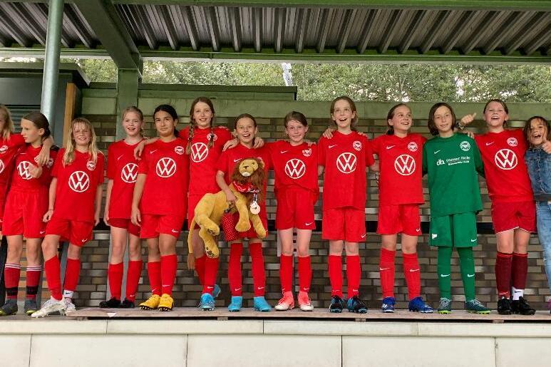 M&auml;dchenfu&szlig;ballmannschaft in roten Trikots mit einem Maskottchen auf einer Trib&uuml;ne, l&auml;chelnd f&uuml;r das Foto aufgestellt.