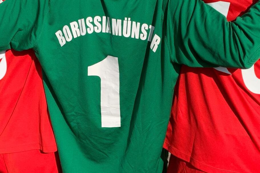 Nahaufnahme von drei Fu&szlig;ballspielern: einer in gr&uuml;nem Trikot mit &bdquo;BORUSSIAM&Uuml;NSTER&ldquo; und zwei in roten Trikots.