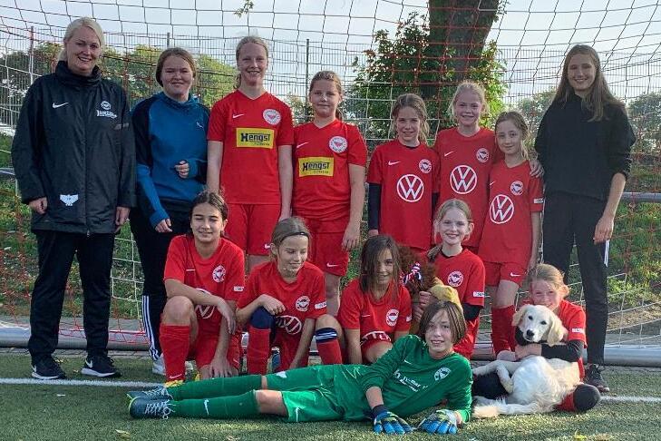 M&auml;dchenfu&szlig;ballmannschaft in roten Trikots posiert mit Trainerin vor einem Fu&szlig;balltor auf einem Kunstrasenplatz.