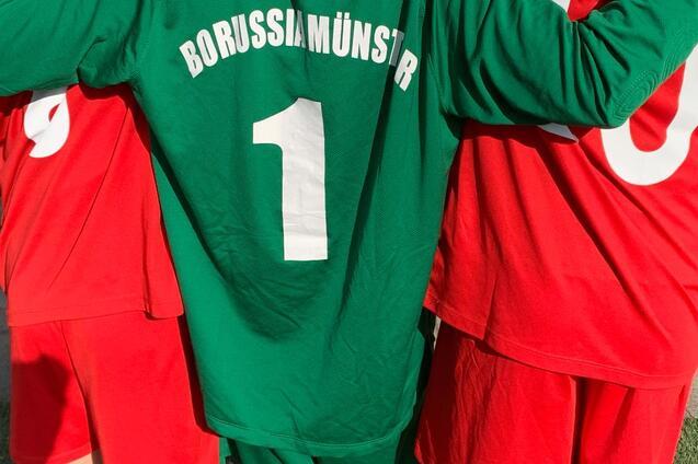 Nahaufnahme von drei Fu&szlig;ballspielern: einer in gr&uuml;nem Trikot mit &bdquo;BORUSSIAM&Uuml;NSTER&ldquo; und zwei in roten Trikots.