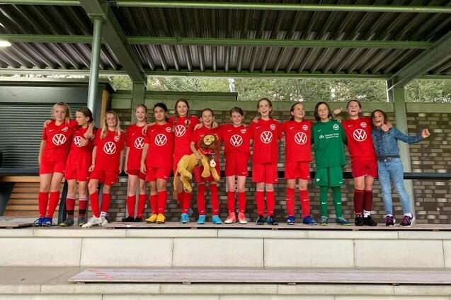 M&auml;dchenfu&szlig;ballmannschaft in roten Trikots mit einem Maskottchen auf einer Trib&uuml;ne, l&auml;chelnd f&uuml;r das Foto aufgestellt.