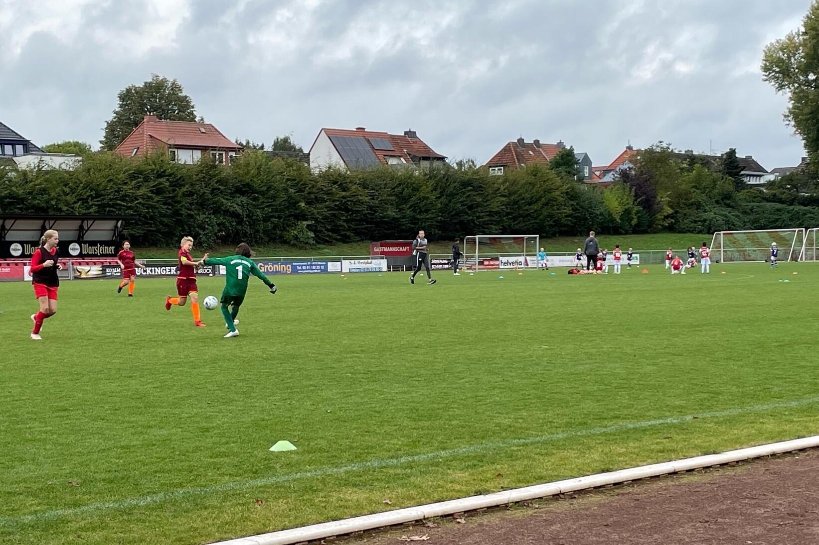 Ein Fu&szlig;ballspiel mit Spielern in roten und gr&uuml;nen Trikots auf einem Rasenplatz, im Hintergrund Tore und Zuschauer.