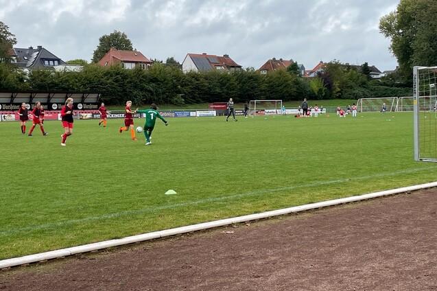 Ein Fu&szlig;ballspiel mit Spielern in roten und gr&uuml;nen Trikots auf einem Rasenplatz, im Hintergrund Tore und Zuschauer.