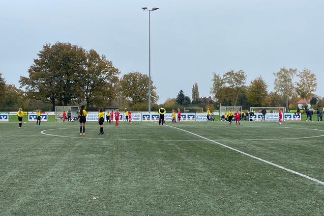 Kindermannschaft auf einem k&uuml;nstlichen Fu&szlig;ballplatz, umgeben von B&auml;umen, spielt unter grauem Himmel.