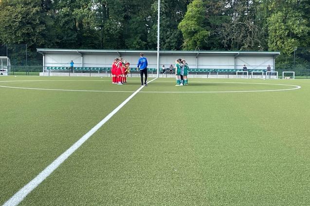 Zwei Fu&szlig;ballmannschaften in Rot und Gr&uuml;n stehen auf einem Kunstrasenplatz im Kreis, eingeladet von einem Trainer.