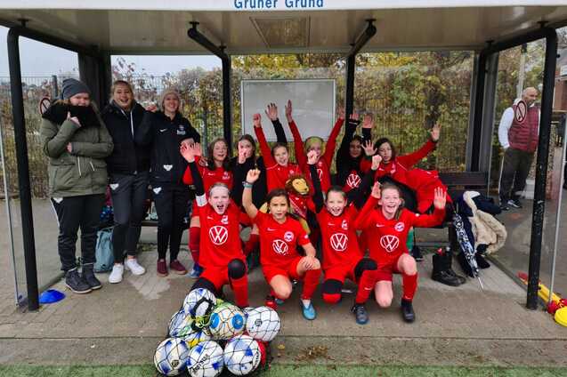 Mannschaft von jungen Fu&szlig;ballspielern in roten Trikots, die fr&ouml;hlich auf einer Bank stehen, umgeben von B&auml;llen und Betreuern.