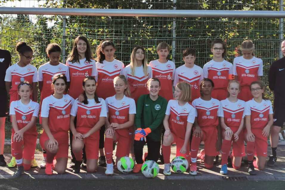 M&auml;dchen-Fu&szlig;ballmannschaft mit Trainer steht vor einem Tor auf einem Sportplatz, bunte B&auml;lle liegen im Vordergrund.