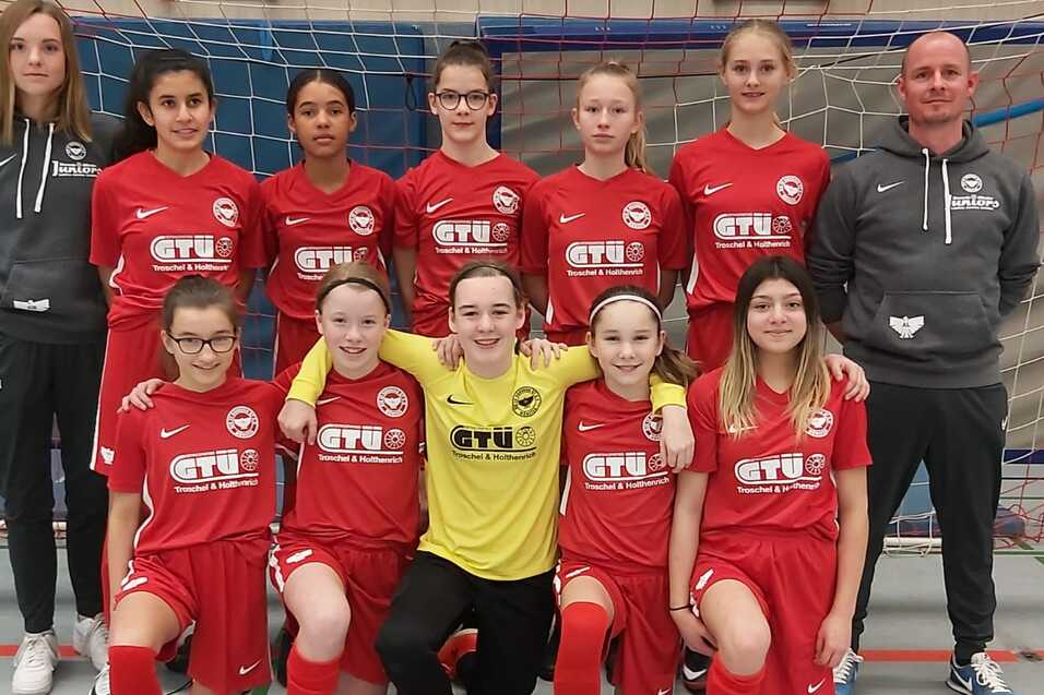 Gruppe von zw&ouml;lf M&auml;dchen in roten Fu&szlig;balltrikots mit Trainer, posierend vor einem Fu&szlig;balltor in einer Sporthalle.