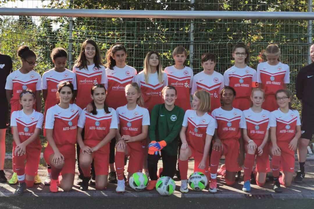 M&auml;dchen-Fu&szlig;ballmannschaft mit Trainer steht vor einem Tor auf einem Sportplatz, bunte B&auml;lle liegen im Vordergrund.