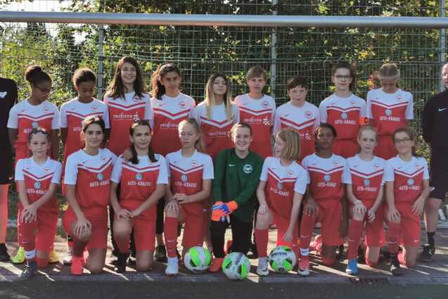 M&auml;dchen-Fu&szlig;ballmannschaft mit Trainer steht vor einem Tor auf einem Sportplatz, bunte B&auml;lle liegen im Vordergrund.
