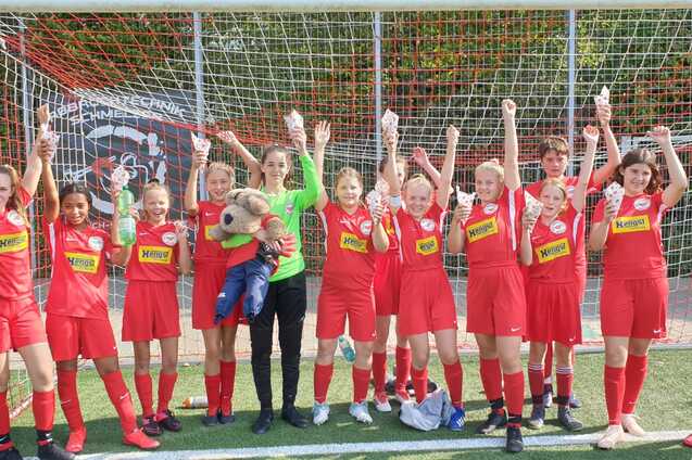 M&auml;dchenfu&szlig;ballmannschaft in roten Trikots feiert vor einem Fu&szlig;balltor mit H&auml;nden in der Luft.