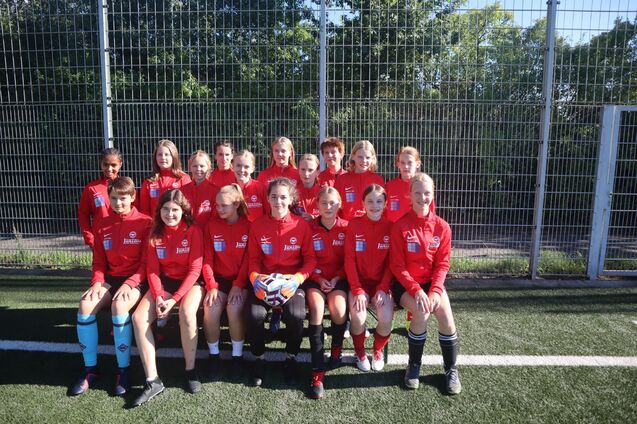 Gruppenfoto eines Fu&szlig;ballteams in roten Trikots auf einem Kunstrasenplatz, mit einer Torfrau und einem Ball im Vordergrund.
