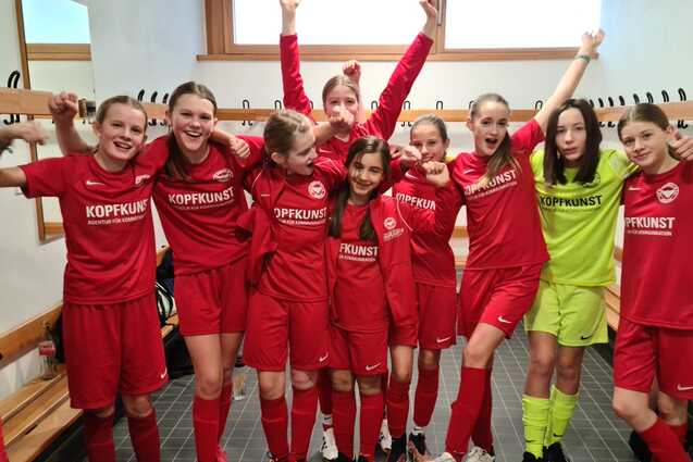 M&auml;dchenmannschaft in roten Fu&szlig;balltrikots jubelt in Umkleidekabine, einige halten Arme hoch, mit einem Torwart in Gelb.