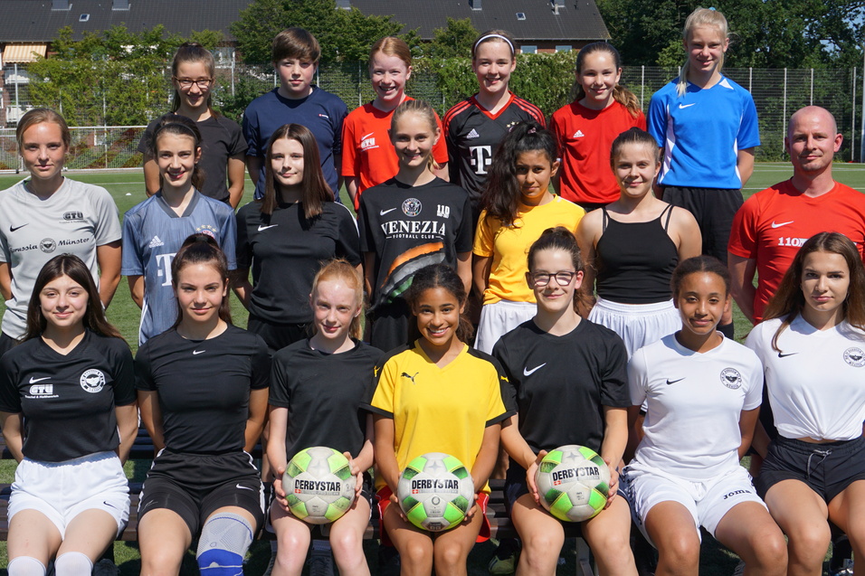 Gruppenfoto von 17 jungen Fu&szlig;ballspielerinnen in Sportkleidung, zusammen mit einem Trainer auf einem Fu&szlig;ballplatz.