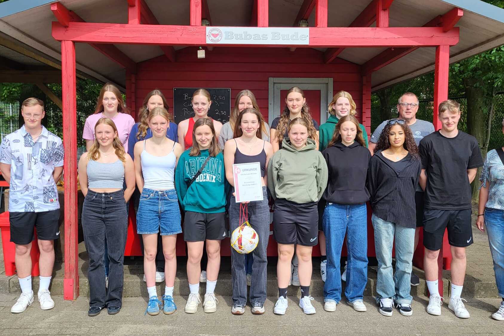 Gruppenfoto von 16 Personen vor einer roten Holzh&uuml;tte, einige halten einen Volleyball, alle l&auml;cheln in die Kamera.