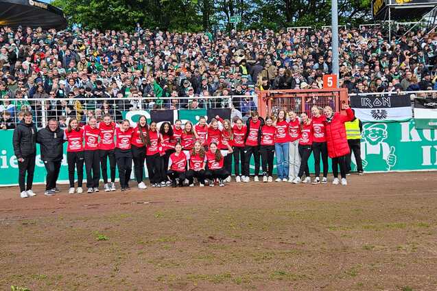 Gruppe von Sportlerinnen in roten Shirts posiert vor jubelndem Publikum im Stadion.