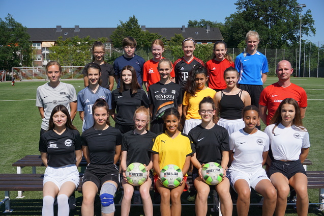 Gruppenfoto von 17 jungen Fu&szlig;ballspielerinnen in Sportkleidung, zusammen mit einem Trainer auf einem Fu&szlig;ballplatz.