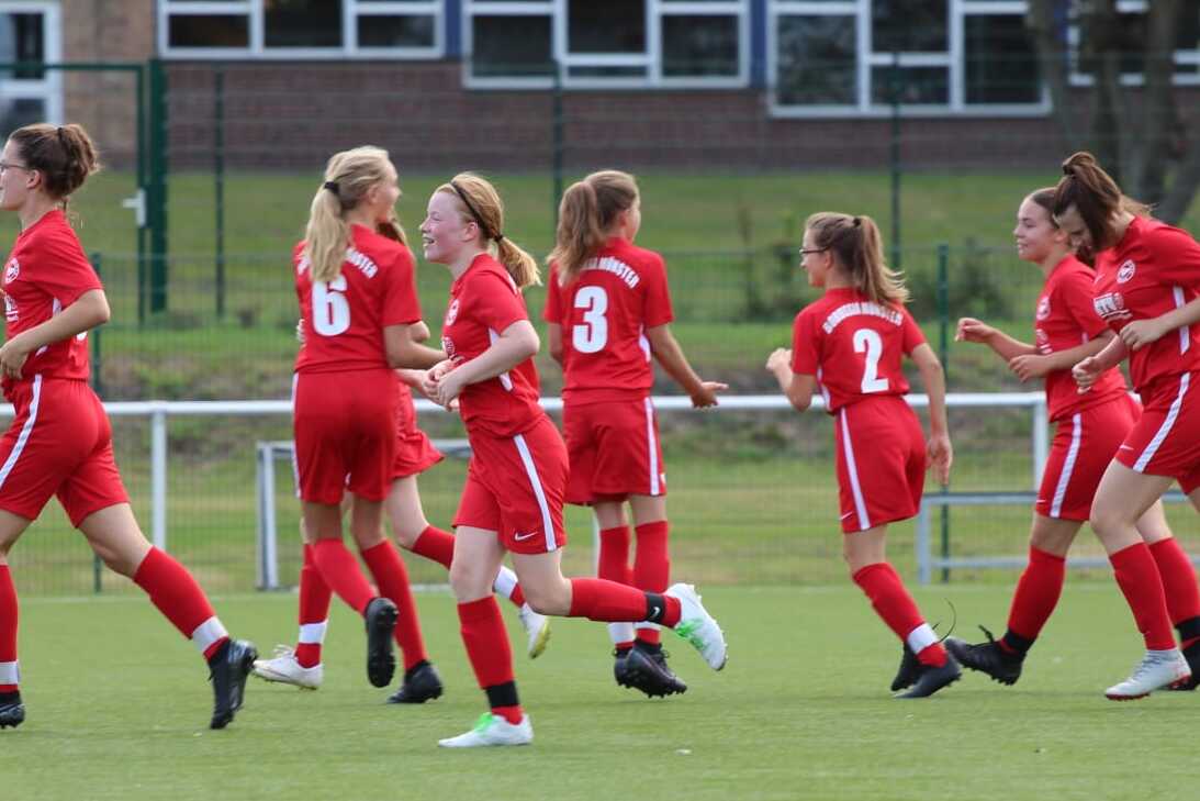 Gruppe junger M&auml;dchen in roten Fu&szlig;balltrikots jubelt auf einem gr&uuml;nen Fu&szlig;ballfeld nach einem Tor.