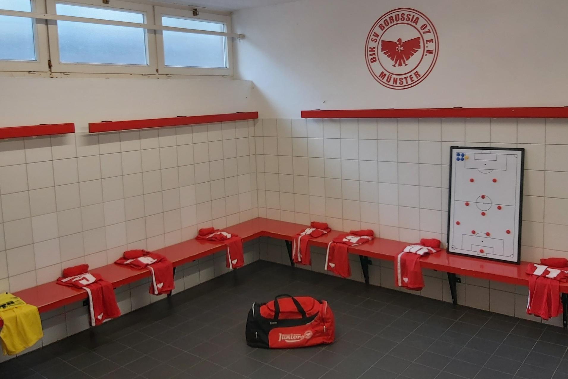 Umkleideraum mit roten B&auml;nken, Spielertrikots, einer Sporttasche und einem Strategieboard an der Wand.