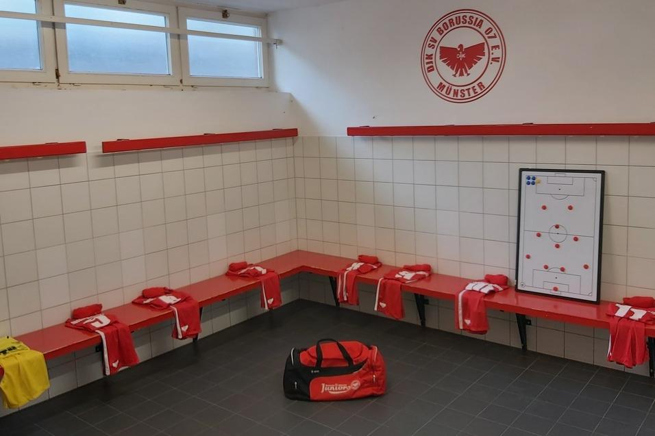 Umkleideraum mit roten B&auml;nken, Spielertrikots, einer Sporttasche und einem Strategieboard an der Wand.