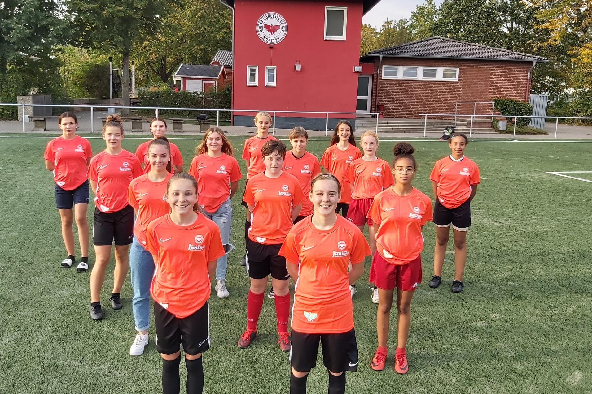 Gruppe von 14 M&auml;dchen in roten Fu&szlig;balltrikots steht auf einem Fu&szlig;ballfeld vor einem Vereinsgeb&auml;ude.