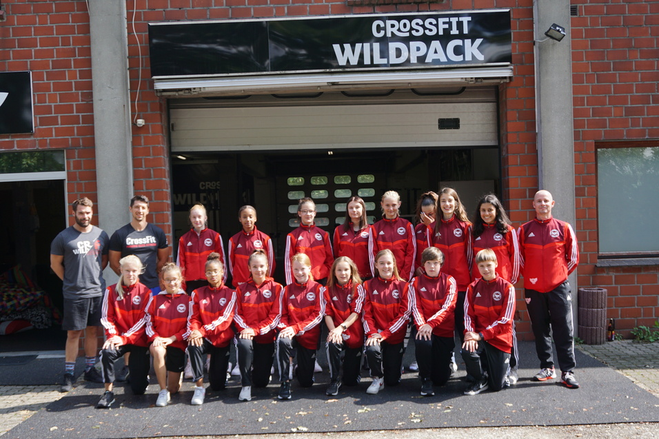 Gruppe von 15 Sportlern in roten Trainingsanz&uuml;gen vor dem Eingang von CrossFit Wildpack, umgeben von Ziegelbauten.