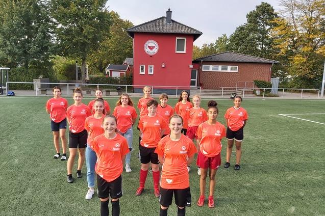 Gruppe von 14 M&auml;dchen in roten Fu&szlig;balltrikots steht auf einem Fu&szlig;ballfeld vor einem Vereinsgeb&auml;ude.