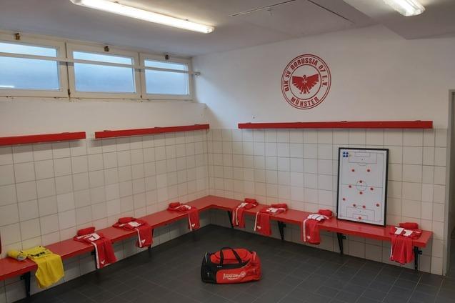 Umkleideraum mit roten B&auml;nken, Spielertrikots, einer Sporttasche und einem Strategieboard an der Wand.